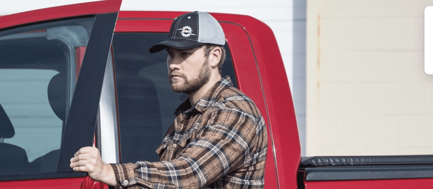 A man wearing an AMSOIL hat stands behind a truck, showcasing the high-quality headwear available in the AMSOIL free hat promotion.