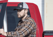 A man wearing an AMSOIL hat stands behind a truck, showcasing the high-quality headwear available in the AMSOIL free hat promotion.