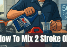 Mechanic pouring AMSOIL 2-stroke oil into a measuring cup beside a red gas can in a garage, with a dirt bike and chainsaw in the background. A ratio chart shows 50:1 equals 2.6 oz per gallon.